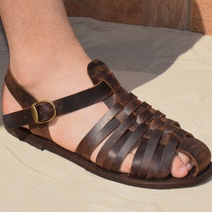 May include: Brown leather sandal with woven straps and a buckle closure. The sandal features a flat sole and a strap around the ankle. The sandal is designed for comfort and style.