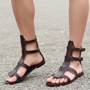 May include: Brown leather gladiator sandals with multiple straps and buckles. The sandals have a high ankle design and a flat sole. The footwear is shown on a person's feet, with a gravel background.