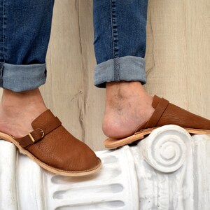 May include: Brown leather slip-on shoes with a buckle strap. The shoes are worn by a person with rolled up blue jeans.
