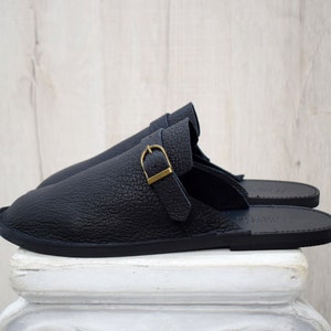 May include: A pair of black leather slippers with a textured finish. Each slipper features a strap with a gold-colored buckle. The slippers are displayed on a white pedestal against a light wood background.