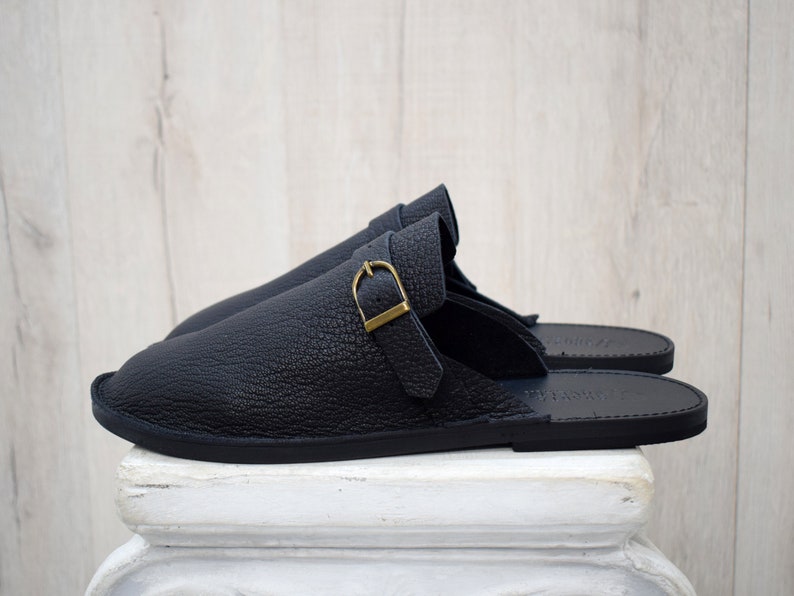May include: A pair of black leather slippers with a textured finish. Each slipper features a strap with a gold-colored buckle. The slippers are displayed on a white pedestal against a light wood background.