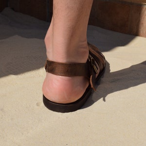 May include: Brown leather sandal with multiple straps and a buckle closure. The sandal has a dark brown sole and a strap around the ankle. The image shows the back of the foot and the sandal on a light-colored surface.