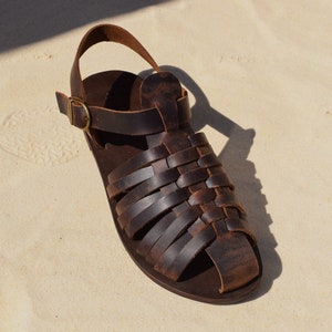 May include: A single brown leather sandal with a woven design across the top. The sandal has a strap around the ankle and a buckle closure. The sandal is sitting on a light-colored surface.