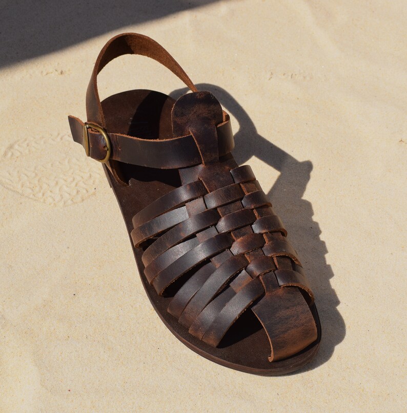 May include: A single brown leather sandal with a woven design across the top. The sandal has a strap around the ankle and a buckle closure. The sandal is sitting on a light-colored surface.