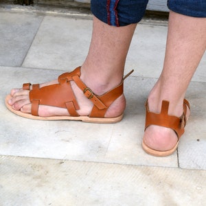 K&ouml;nnte beinhalten: Ein Paar braune Ledersandalen mit Schnallen. Die Sandalen haben einen Riemen, der um den Kn&ouml;chel geht, und einen Zehenriemen.