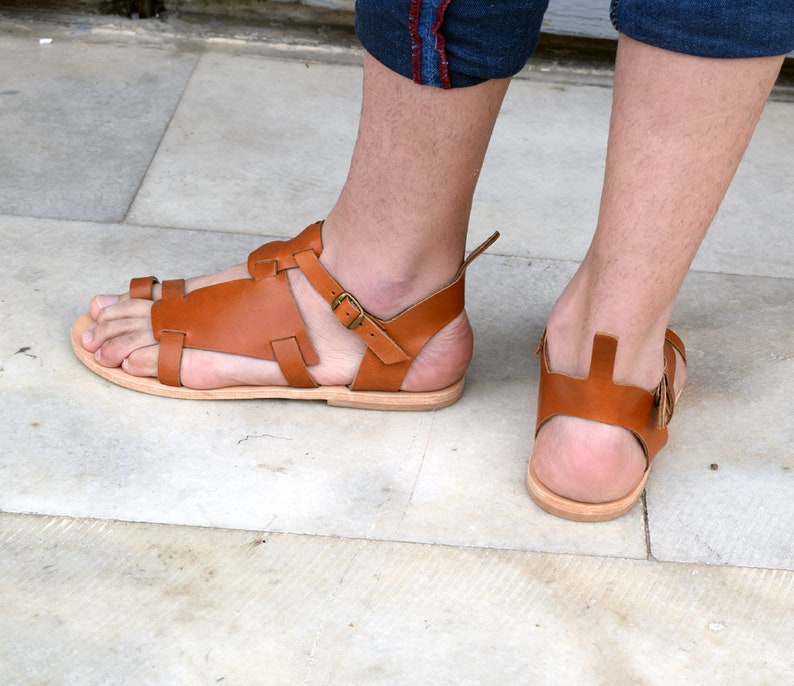 K&ouml;nnte beinhalten: Ein Paar braune Ledersandalen mit Schnallen. Die Sandalen haben einen Riemen, der um den Kn&ouml;chel geht, und einen Zehenriemen.