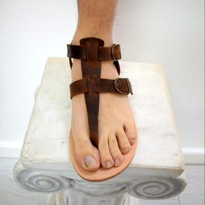 Puede incluir: Sandalias de cuero marr&oacute;n con correas ajustables y cierres de hebilla. Las sandalias tienen un dise&ntilde;o sencillo con una correa para el dedo del pie tipo tanga. La plantilla es de color beige claro. Las sandalias est&aacute;n sobre una superficie blanca.