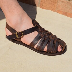 May include: Brown leather sandals with a woven design across the foot and a buckle closure. The sandals have a strap around the ankle and are shown on a sandy surface. The sandals are a size 9.