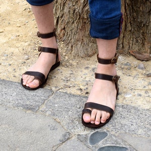 May include: A pair of brown leather sandals with buckle closures. The sandals have a simple design and are worn on a person's feet.