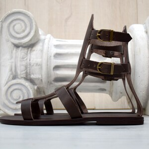 May include: Brown leather gladiator sandals with multiple straps and buckle closures. The sandals have a flat sole and a high, supportive back. The buckles are gold-colored. The sandals are displayed in front of a white, classical-style column.