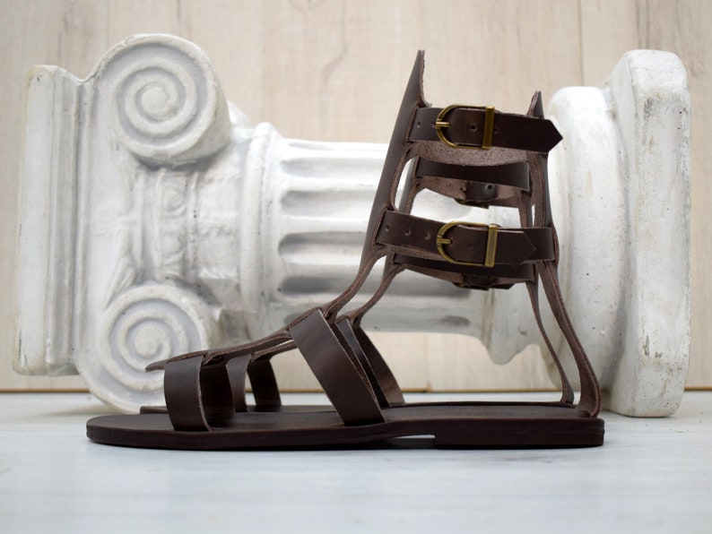 May include: Brown leather gladiator sandals with multiple straps and buckle closures. The sandals have a flat sole and a high, supportive back. The buckles are gold-colored. The sandals are displayed in front of a white, classical-style column.