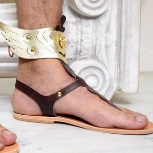 May include: A brown leather sandal featuring a gold-colored metallic ankle cuff with a prominent wing design on the side. The sandal includes a flat tan sole, thin brown leather straps, and a small circular gold-toned emblem on the ankle piece.