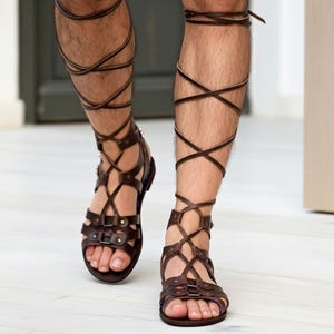 Puede incluir: Sandalias de gladiador en cuero marr&oacute;n oscuro con m&uacute;ltiples tiras horizontales sobre el pie y cordones largos que se envuelven alrededor de la parte inferior de la pierna. Incluyen remaches met&aacute;licos y una suela plana para un ajuste seguro.