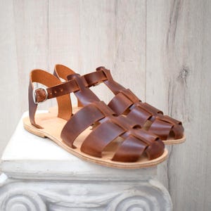 May include: A pair of brown leather sandals with a cage-like design. The sandals have a buckle closure and a light-colored sole. The sandals are displayed on a white pedestal against a light wood-grain background.