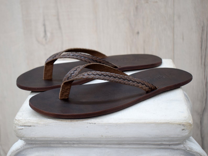 Op de afbeelding: Een paar bruine leren teenslippers met gevlochten bandjes. De sandalen worden gepresenteerd op een wit oppervlak, wat hun eenvoudige ontwerp en natuurlijke materialen benadrukt. De teenslippers zijn donkerbruin.
