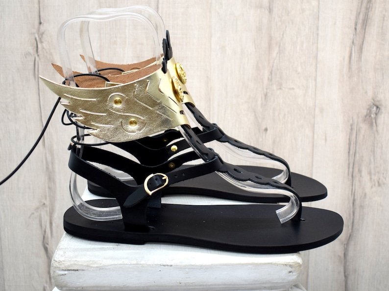 May include: A pair of black flat sandals featuring gold-colored metallic wing attachments at the ankle. The sandals have a thong-style front strap with a scalloped edge, thin black ankle straps with a gold-toned metal buckle, and black soles.