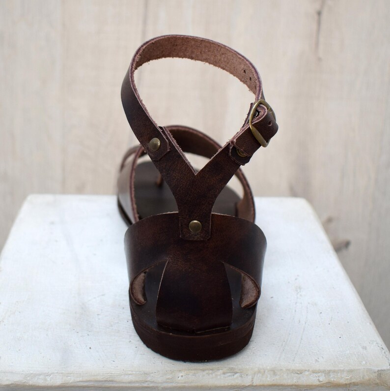 May include: A brown leather sandal with an ankle strap and buckle closure. The sandal features a toe loop and a series of straps across the foot. The sole is a dark brown color, and the sandal appears to be handmade.