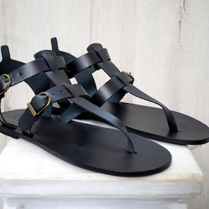 Puede incluir: Un par de sandalias de cuero negro con dise&ntilde;o de correa en T. Las sandalias presentan m&uacute;ltiples correas con hebillas ajustables en tono dorado. Las sandalias se exhiben sobre una superficie blanca contra un fondo de madera clara.