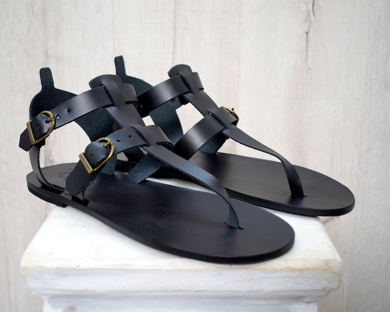 Puede incluir: Un par de sandalias de cuero negro con dise&ntilde;o de correa en T. Las sandalias presentan m&uacute;ltiples correas con hebillas ajustables en tono dorado. Las sandalias se exhiben sobre una superficie blanca contra un fondo de madera clara.