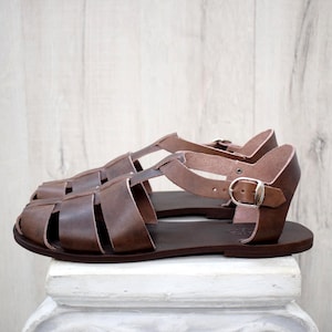 May include: Brown leather sandals with a cage-like design. The sandals have multiple straps and a buckle closure. The soles are a darker shade of brown. The sandals are displayed on a white surface against a light wood background.