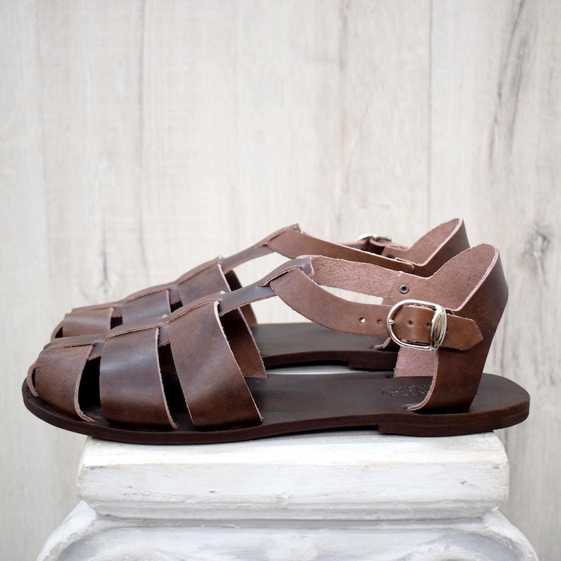 May include: Brown leather sandals with a cage-like design. The sandals have multiple straps and a buckle closure. The soles are a darker shade of brown. The sandals are displayed on a white surface against a light wood background.