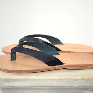 Peut inclure: Une paire de tongs en cuir bleu marine avec des semelles marron clair. Les sandales ont un design simple avec une bride de type lani&egrave;re et une bride qui fait le tour de la cheville. Les sandales sont pos&eacute;es sur une surface blanche.