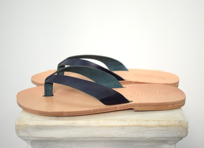 Peut inclure: Une paire de tongs en cuir bleu marine avec des semelles marron clair. Les sandales ont un design simple avec une bride de type lani&egrave;re et une bride qui fait le tour de la cheville. Les sandales sont pos&eacute;es sur une surface blanche.