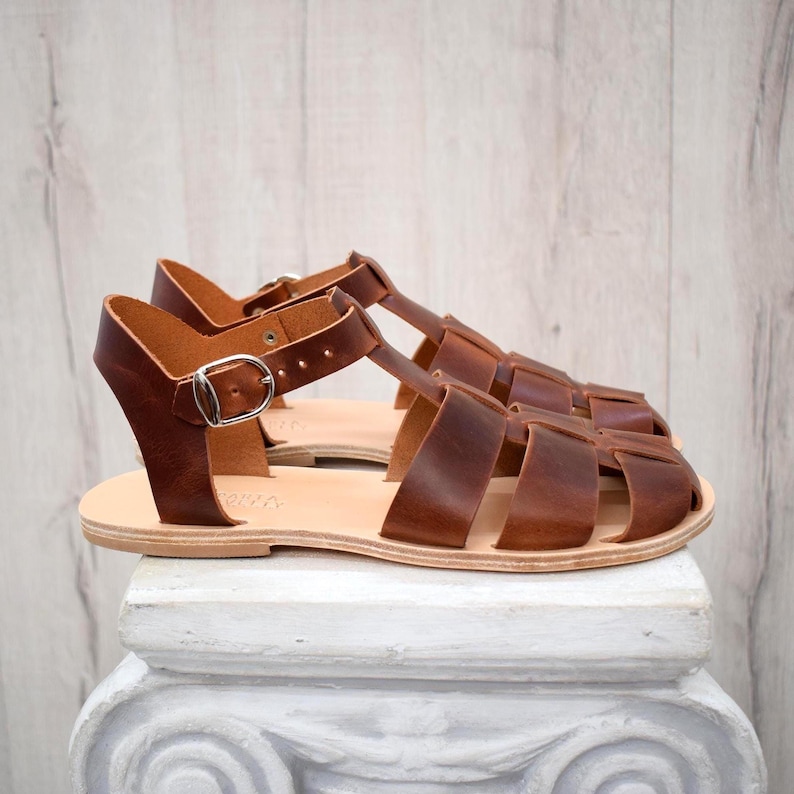 May include: A pair of brown leather sandals with a cage-like design. The sandals have a light-colored sole and a silver buckle closure. The sandals are displayed on a white pedestal against a light wood background.