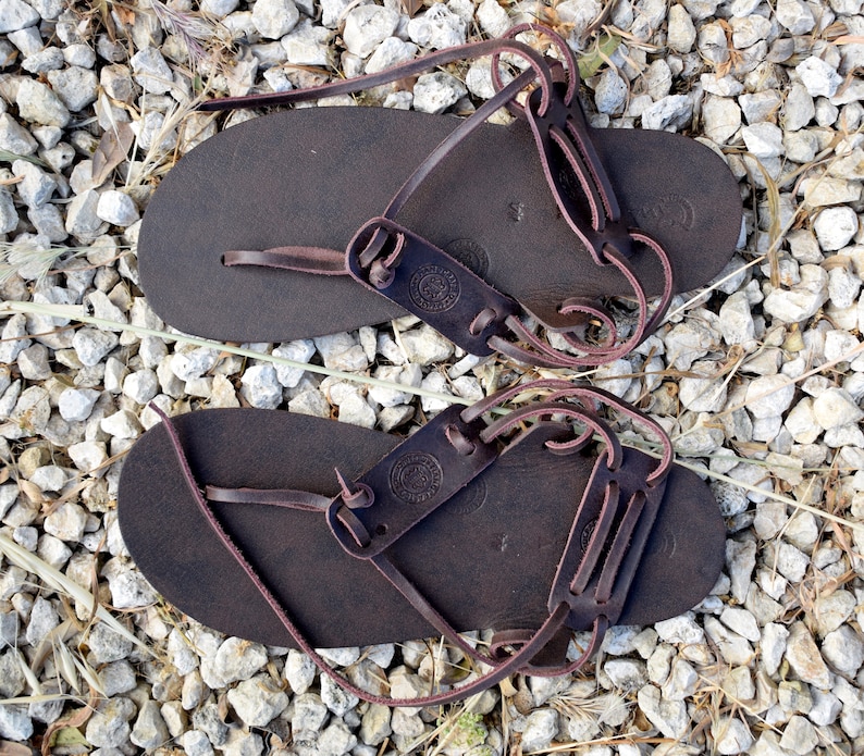 May include: A pair of brown leather sandals with a simple design and adjustable straps. The sandals have a circular logo on the straps.