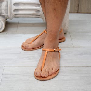 Peut inclure: Sandales en cuir orange avec une bride en T et des lanières de cheville avec boucles. Les sandales ont une semelle plate. Elles sont portées sur un sol en bois clair.