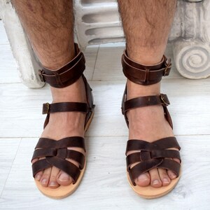 May include: Brown leather sandals with multiple straps across the foot and ankle straps with buckles. The sandals have a light-colored sole. The image shows the sandals on a person's feet.