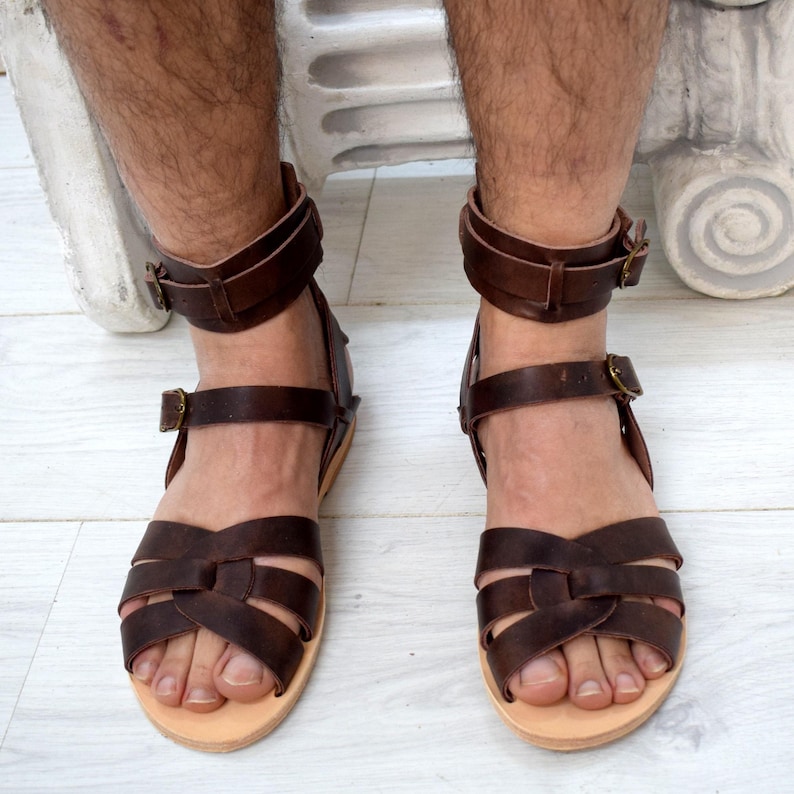 May include: Brown leather sandals with multiple straps across the foot and ankle straps with buckles. The sandals have a light-colored sole. The image shows the sandals on a person's feet.