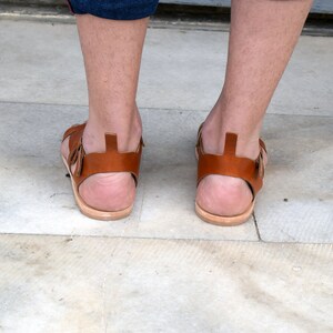 K&ouml;nnte beinhalten: Ein Paar braune Ledersandalen mit einem Riemen &uuml;ber dem Fu&szlig; und einer Schnalle am Kn&ouml;chel.