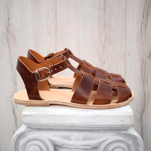 May include: A pair of brown leather sandals with a cage-like design. The sandals have a light-colored sole and a silver buckle closure. The leather appears to be distressed, giving them a vintage look. The sandals are displayed on a white pedestal.