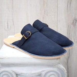 May include: A pair of navy blue leather slippers with a buckle detail. The open-back design features a light tan insole and a light blue trim. The slippers are displayed on a white pedestal against a light wood background.
