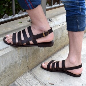 May include: Brown leather sandals with multiple straps and a buckle fastening. The sandals have a flat sole and are being worn.