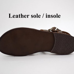 May include: A brown leather sandal with a leather sole and insole. The sandal features a buckle closure and a textured rubber heel. The text "Leather sole / insole" is visible.