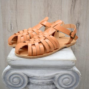 May include: A pair of light brown leather sandals with a woven design. The sandals have a strap around the ankle with a buckle closure. The sandals are displayed on a white, decorative pedestal.