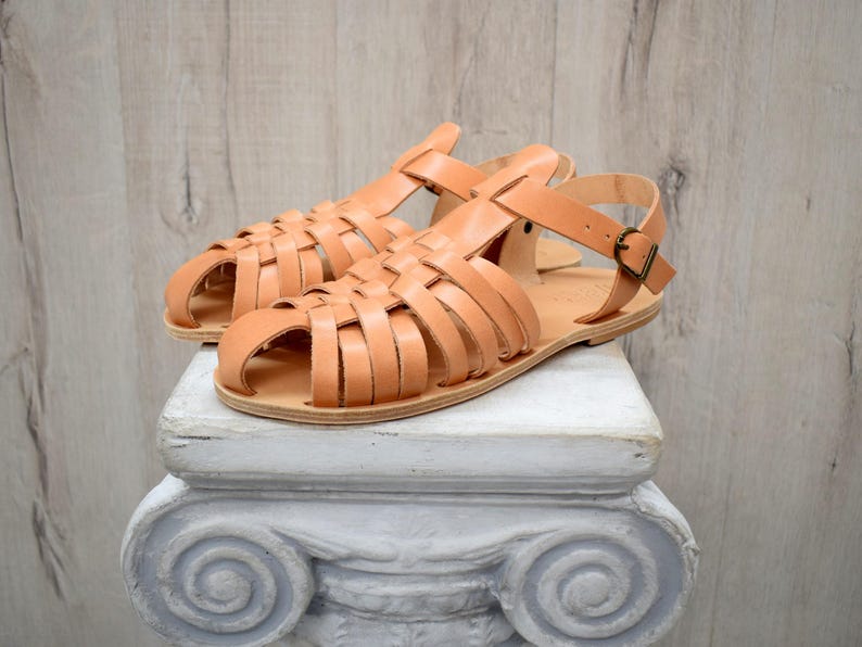 May include: A pair of light brown leather sandals with a woven design. The sandals have a strap around the ankle with a buckle closure. The sandals are displayed on a white, decorative pedestal.