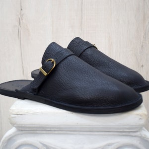 May include: A pair of black leather sandals with a textured finish. Each sandal features a strap with a gold-colored buckle. The sandals are displayed on a white pedestal against a light wood-grain background.