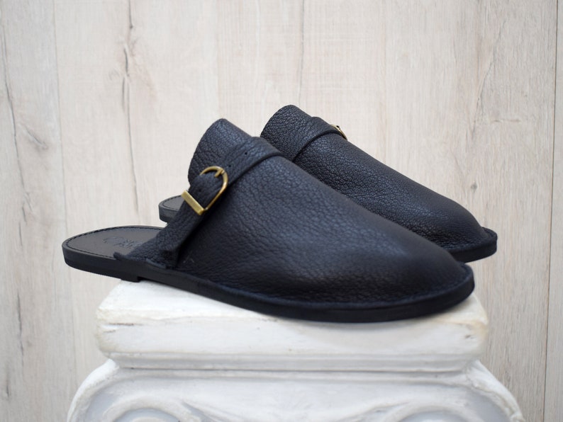 May include: A pair of black leather sandals with a textured finish. Each sandal features a strap with a gold-colored buckle. The sandals are displayed on a white pedestal against a light wood-grain background.
