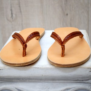 Op de afbeelding: Een paar leren sandalen met een lichtbruin voetbed en een donkerbruin, geweven bandje. De sandalen worden getoond op een wit oppervlak tegen een lichte houten achtergrond. De sandalen zijn een klassieke teenslipper stijl.