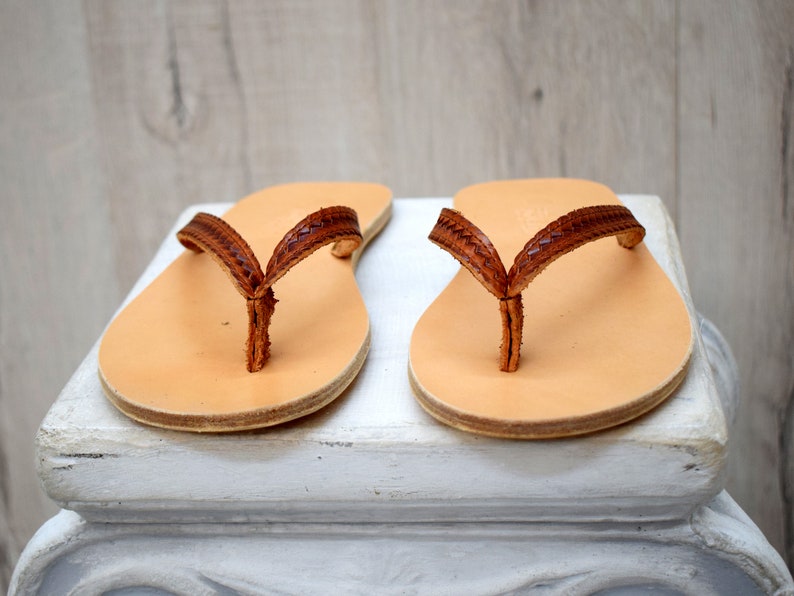 Op de afbeelding: Een paar leren sandalen met een lichtbruin voetbed en een donkerbruin, geweven bandje. De sandalen worden getoond op een wit oppervlak tegen een lichte houten achtergrond. De sandalen zijn een klassieke teenslipper stijl.