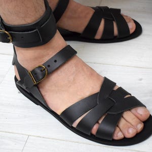 May include: Black leather sandals with multiple straps and ankle straps. The sandals have a flat sole and are secured with adjustable buckles. The design features a classic gladiator style, suitable for warm weather.