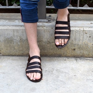 May include: Brown leather sandals with multiple straps. The sandals have a simple design and are worn on a person's feet.
