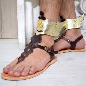 May include: A pair of dark brown leather sandals featuring a flat tan sole and a vertical strap with a scalloped edge. The ankle cuff is made of metallic gold material shaped like wings, accented with a circular gold emblem on the front side.