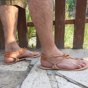 May include: A pair of brown leather sandals with a criss-cross strap design. The sandals are worn on a person's feet, standing on a stone surface.