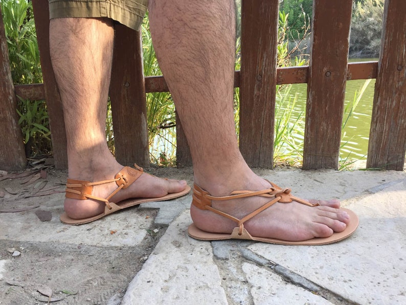 May include: A pair of brown leather sandals with a criss-cross strap design. The sandals are worn on a person's feet, standing on a stone surface.
