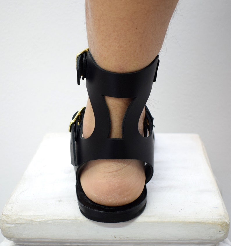 May include: Black leather gladiator sandals with gold-colored buckles. The sandals feature a cutout design on the ankle strap and a flat sole. The image shows the back of the sandal and part of the leg.