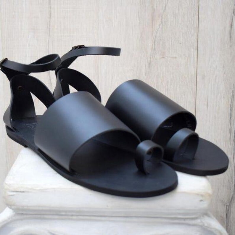 May include: A pair of black leather sandals with a toe loop and ankle strap. The sandals have a minimalist design with a flat sole and are displayed on a white surface.